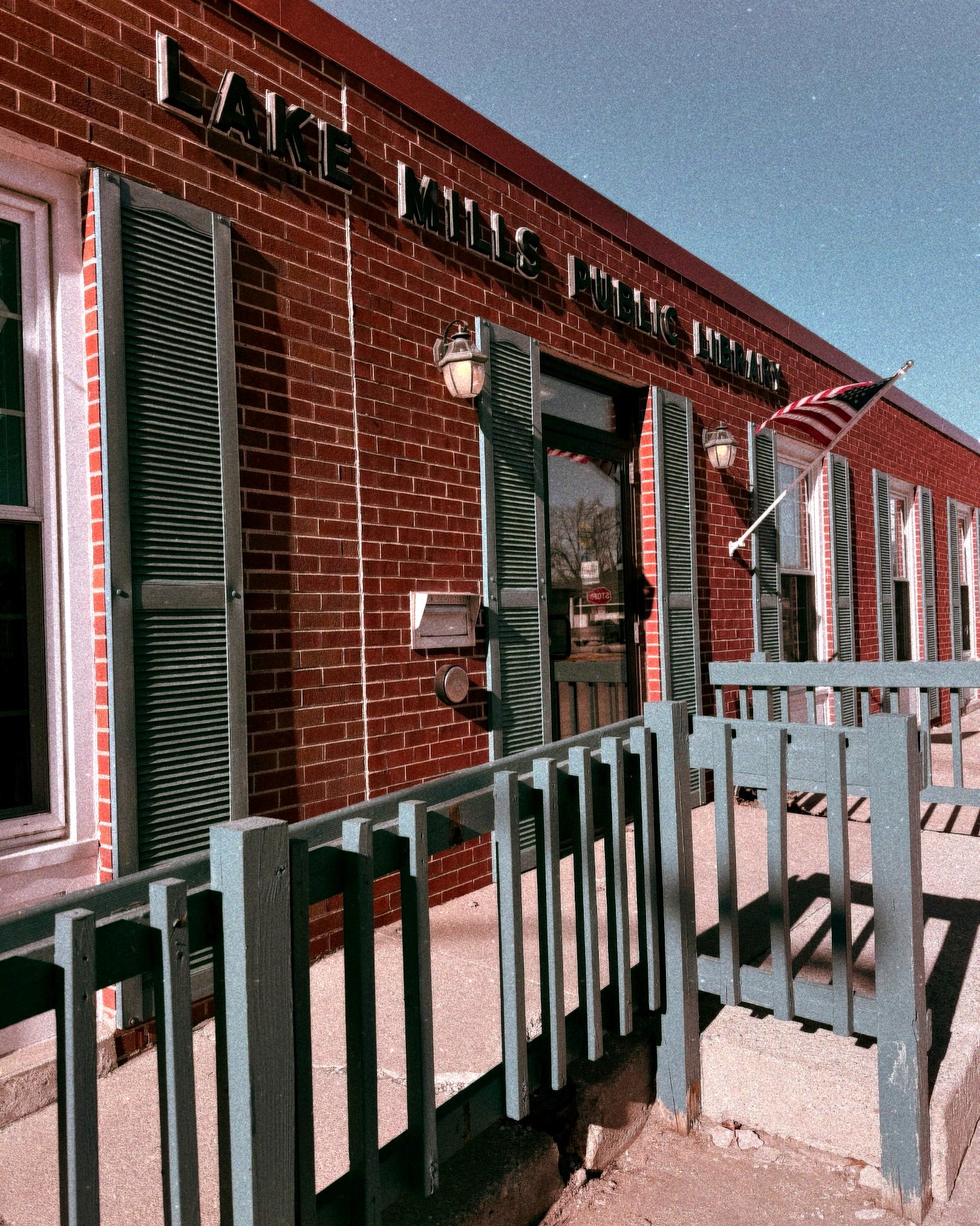 Photo of the Lake Mills Public LIbrary with blue shutters and blue railing on entrance ramp.